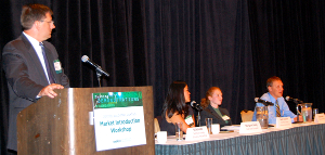 Photo of speakers facing an audience, the speaker at the left standing at a lectern and the other speakers sitting at a table.